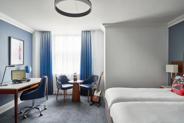 A hotel bedroom with two beds, a desk with a record player, two armchairs at a small round table, and an electric guitar on a stand by the window.