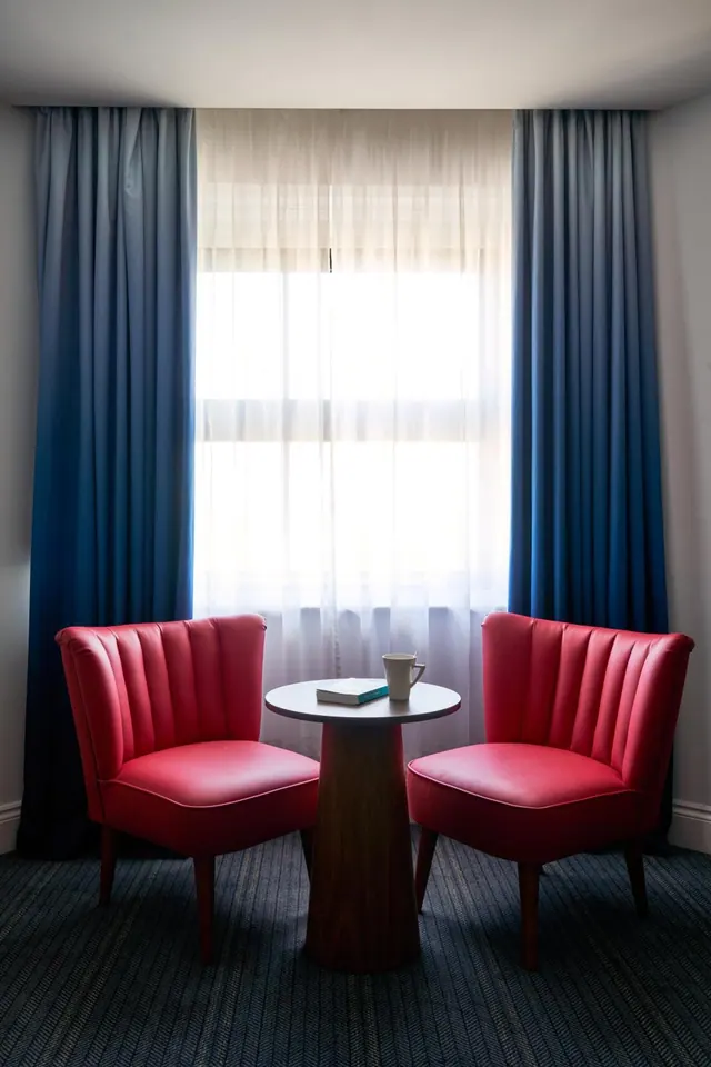 Two red upholstered armchairs flank a small round table holding a mug and a book, in front of a window with sheer white curtains and blue drapes.