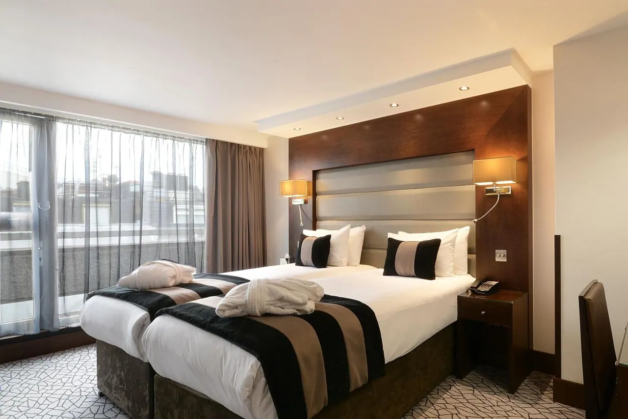 A bedroom with two beds made up with white linen, striped runners and bathrobes, beside floor-to-ceiling windows with sheer curtains that look out onto neighbouring buildings.