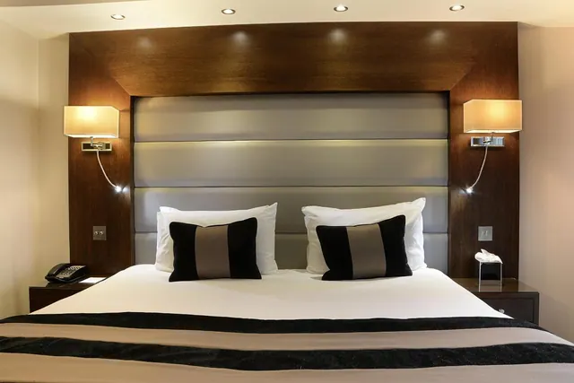 A made-up double bed with white pillows and two black and taupe cushions sits in front of a wood-panelled headboard, with a wall lamp on each side.