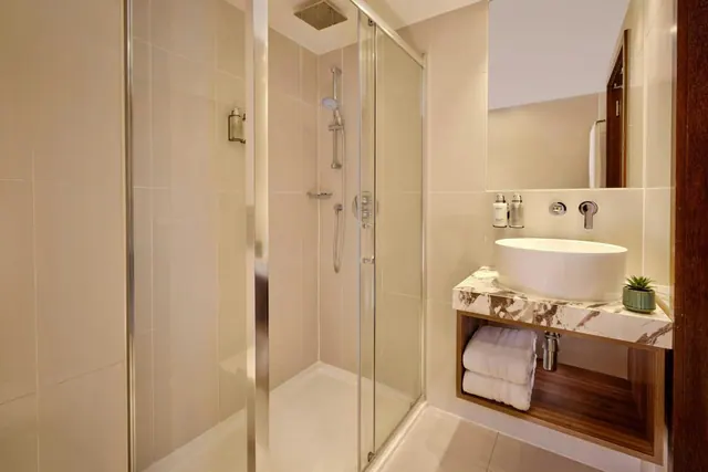 A bathroom with a glass-enclosed walk-in shower, a vessel sink on a marble countertop, and folded towels on an open shelf below the sink.