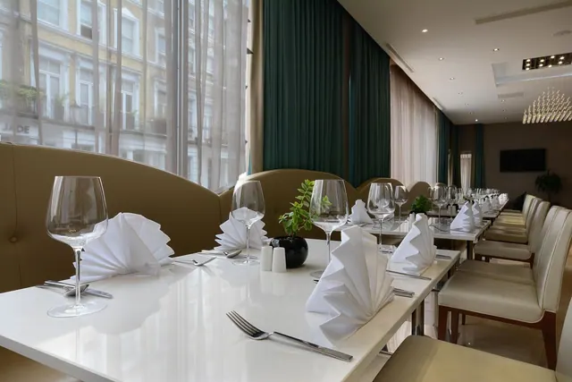 A restaurant dining room with set tables, folded white napkins and wine glasses, with large windows looking onto a street lined with buildings.