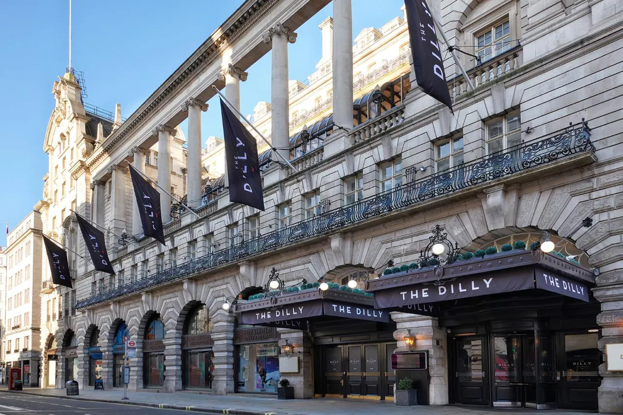 Exterior of The Dilly hotel showing its stone facade, classical columns, black hanging banners and entrance canopies on a sunny street.
