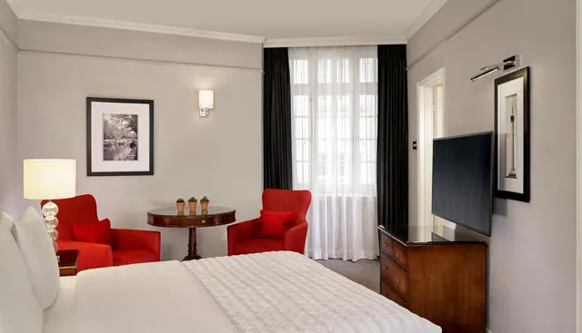 A hotel bedroom with a made bed, a pair of red armchairs beside a small round table, a flat-screen television on a wooden chest of drawers, and sheer-curtained windows.