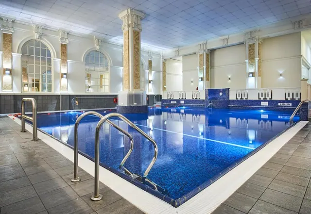 An indoor swimming pool with blue mosaic tiles, steel pool ladders, a lane line, and classical columns and arched windows along the walls.
