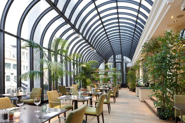 A restaurant dining room with a curved glass roof, rows of tables and chairs, and potted plants along the walls.