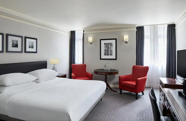 A staged photo of a bedroom with a king bed, two red armchairs beside a small round table, and a wooden desk with a chair and a wall-mounted television.