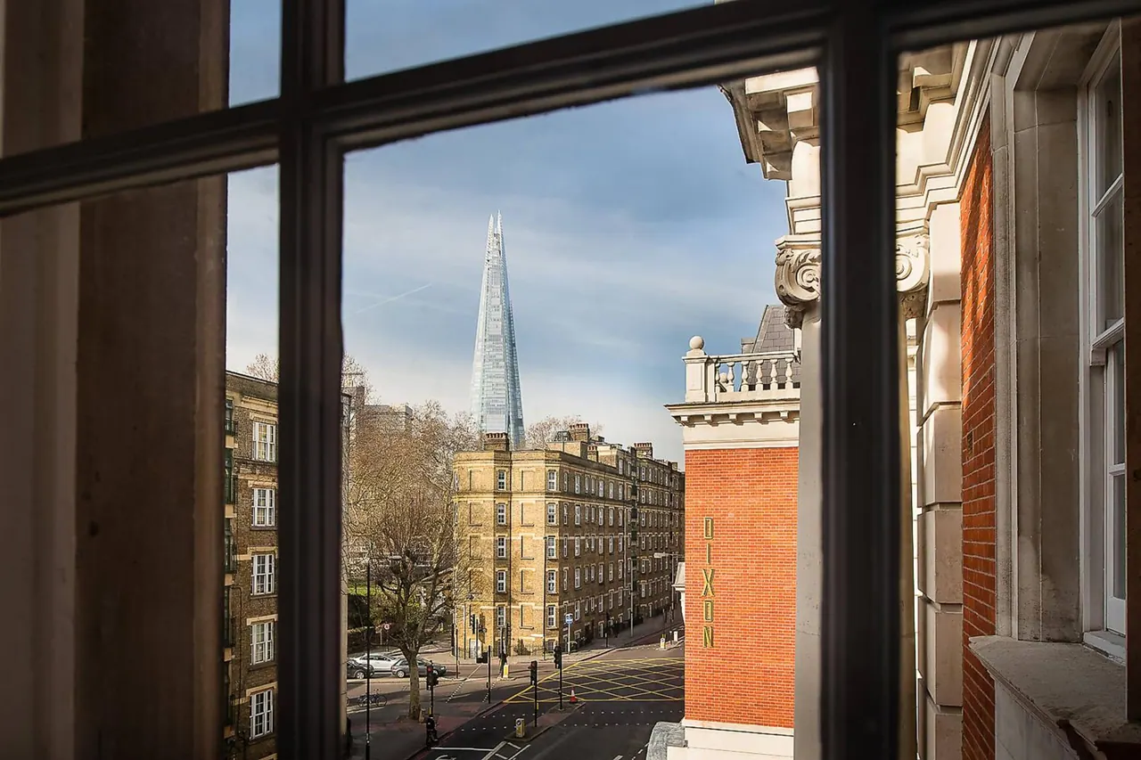 View through a window with glazing bars across a street of stone and brick buildings toward The Shard, with a brick building bearing the name DIXON in the foreground.
