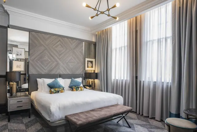 A bedroom with a made king bed, an upholstered bench at its foot, tall curtained windows and a diamond-patterned wood panel behind the headboard.