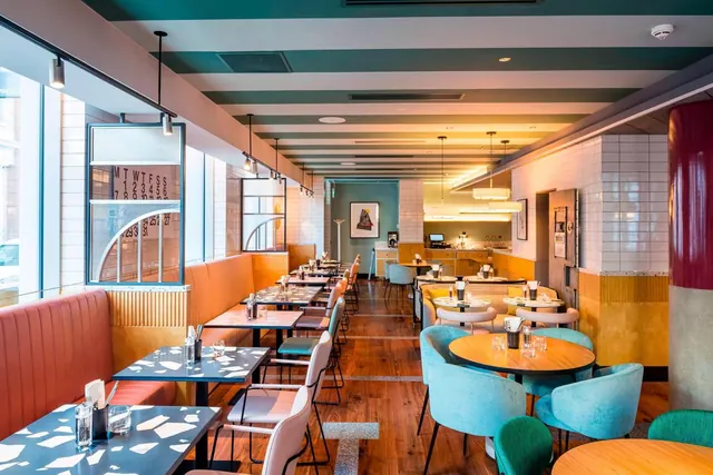 A restaurant dining room with orange banquette seating along the windows, round wooden tables, turquoise chairs and a tiled counter at the back.