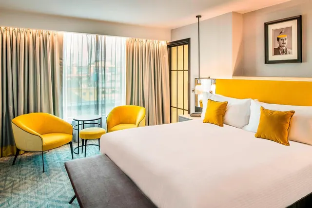 A king bed with yellow cushions faces two yellow armchairs and a small round table beside a window with sheer and lined curtains.
