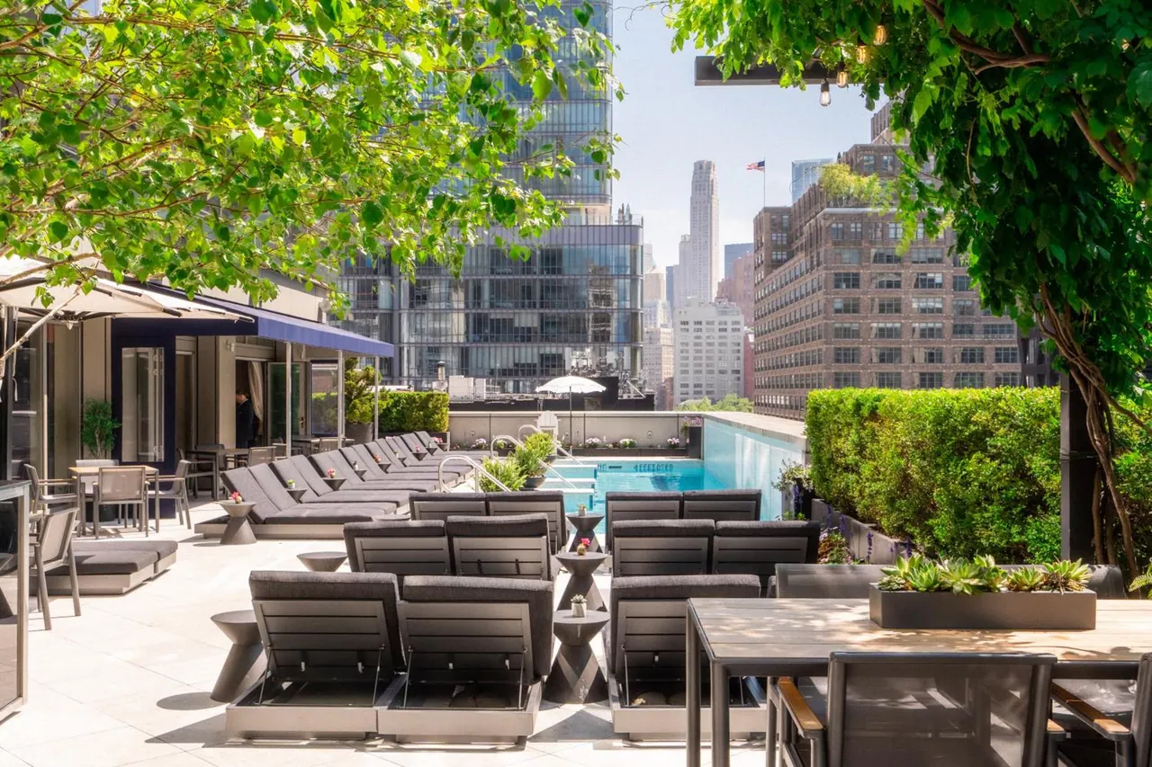 A rooftop pool with grey lounge chairs and dining tables on a stone deck, with high-rise buildings and a flagpole visible behind it.