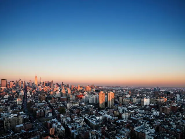 Aerial view over a dense city at dusk with the Empire State Building visible on the skyline under a blue-to-orange sky.
