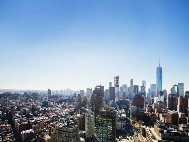 Elevated view over a dense Manhattan skyline toward One World Trade Center under a clear blue sky.