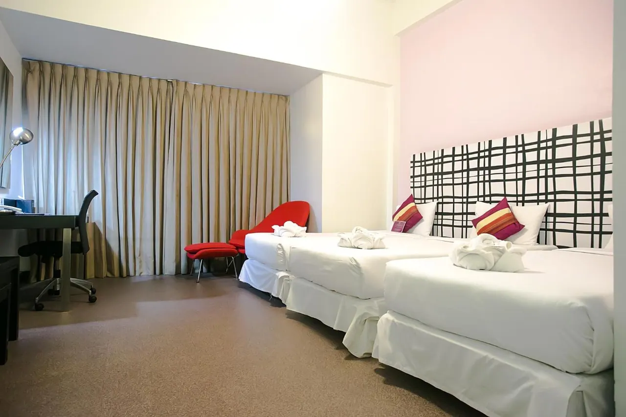 A hotel bedroom with two white beds, a red armchair with ottoman, a desk with chair, and beige curtains covering the window.