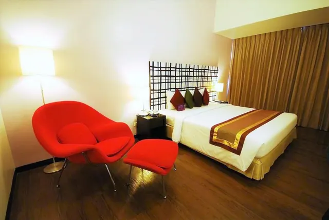 A hotel bedroom with a red armchair and matching footstool beside a made bed with patterned cushions and a brown runner, lit by lamps with closed curtains behind.