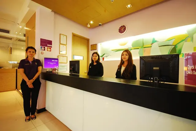 Three staff members stand at a black and white reception counter, with a purple welcome screen, a wall clock and a red check-in and check-out sign nearby.
