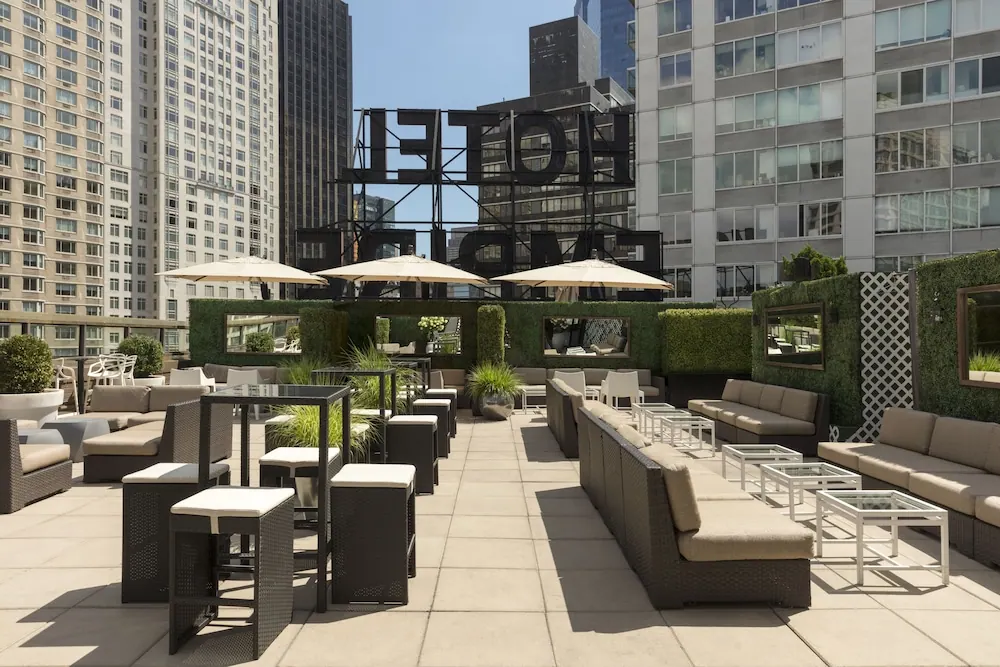A rooftop terrace with wicker lounge seating, high bar tables, cream umbrellas and a large black hotel sign rises above surrounding city buildings.