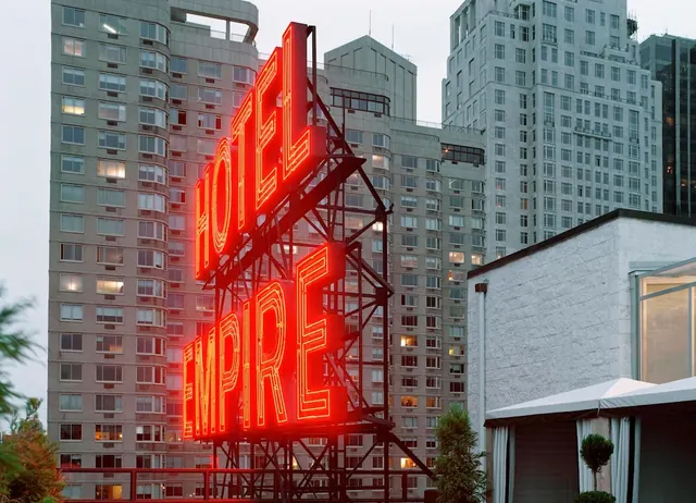 An illuminated red neon Hotel Empire sign on a steel lattice frame, with residential towers and a white rooftop structure behind it at dusk.