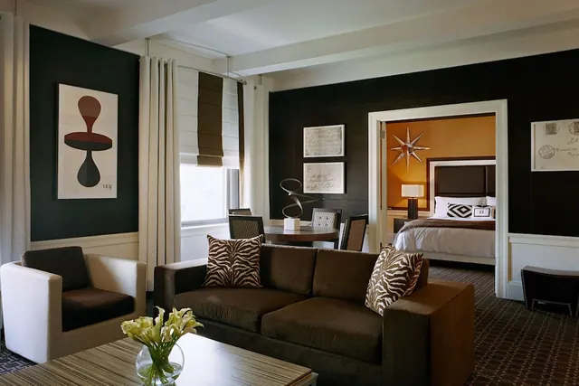 A living area with a brown sofa, white armchair, round dining table and a bedroom visible through an open doorway.