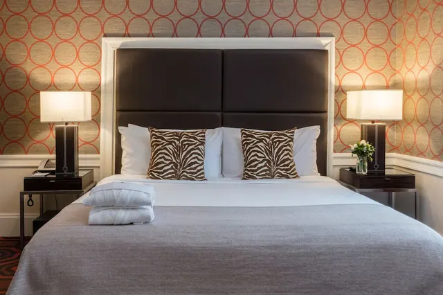 A made-up double bed with a dark leather headboard, zebra-print cushions, two table lamps and folded white bathrobes on the bed.