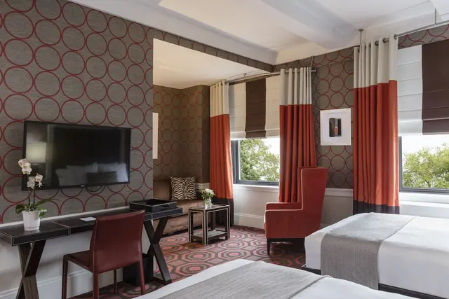 A hotel bedroom with two beds, a wall-mounted television, a desk with a chair, a red armchair and a seating area by windows with trees outside.