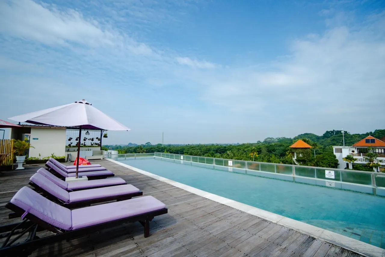 A rooftop swimming pool with purple sun loungers and a white umbrella, with tree-covered land and buildings visible beyond the railing.