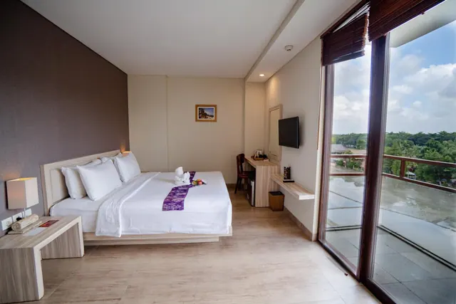 A bedroom with a king bed dressed in white linens and a purple runner, a wall-mounted television and a desk, with sliding glass doors opening onto a balcony with trees and sky beyond.