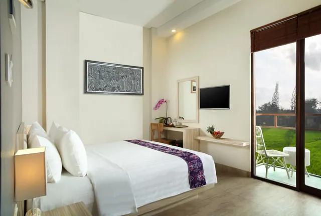A bedroom with a made double bed, a wall-mounted television and a glass door opening onto a balcony with two chairs overlooking green rice fields.