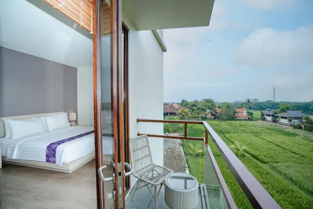 A balcony with wicker chairs and a side table overlooks green rice fields and village rooftops, with an open glass door into a bedroom with a king bed.