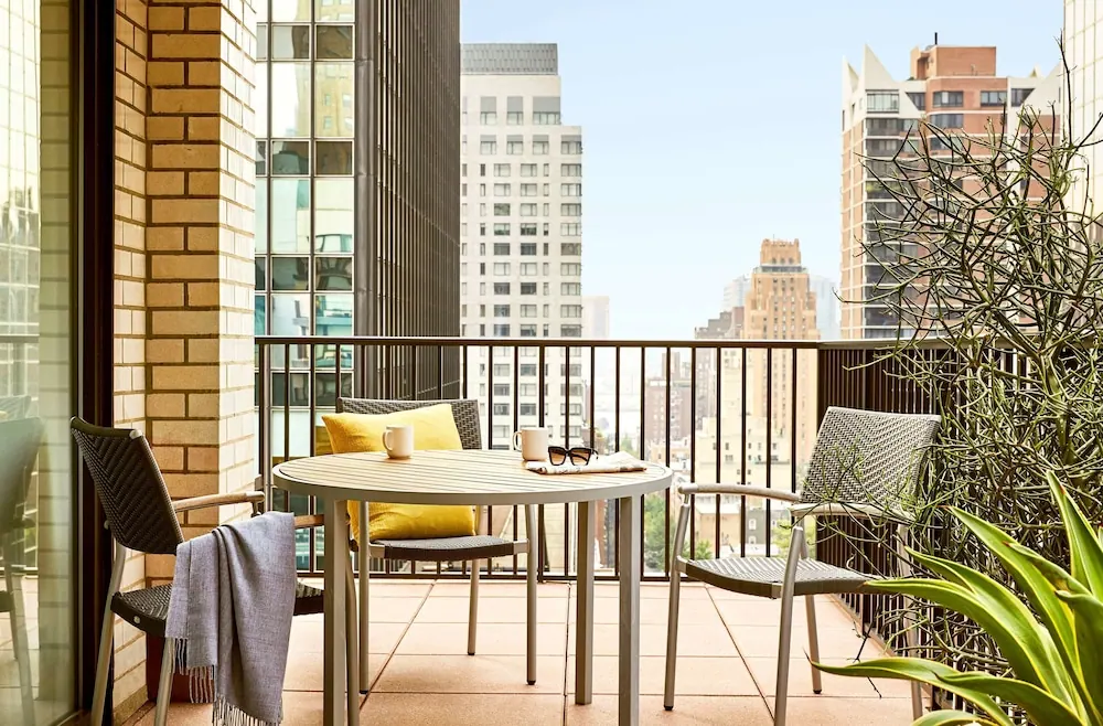A round table with two chairs, a yellow cushion, mugs and sunglasses sits on a tiled balcony overlooking high-rise buildings under a clear sky.