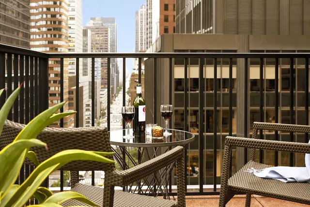 A private balcony with two wicker chairs and a glass table holding a bottle of red wine, two filled glasses and a small dish of snacks, overlooking a city avenue lined with tall buildings.