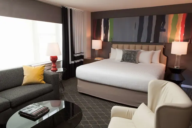A king bed with white linens sits beside a grey sofa with a yellow cushion, a coffee table and a bright window in a hotel bedroom.