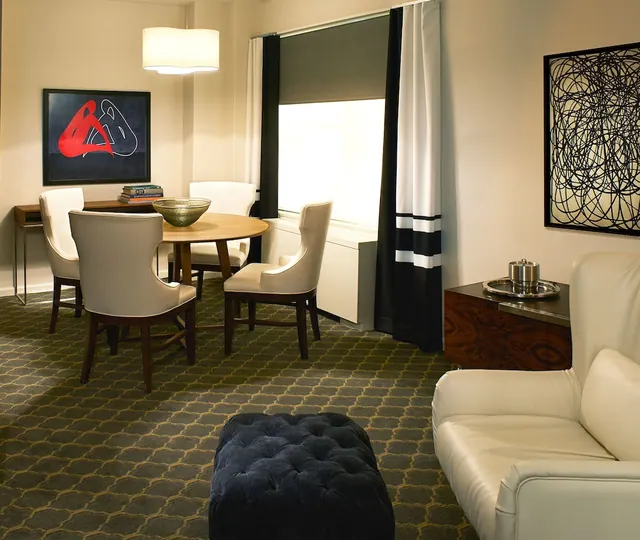 A hotel suite living area with a round wooden table and four upholstered chairs, a white leather sofa, a tufted navy ottoman, a wooden sideboard and a window with black and white curtains.