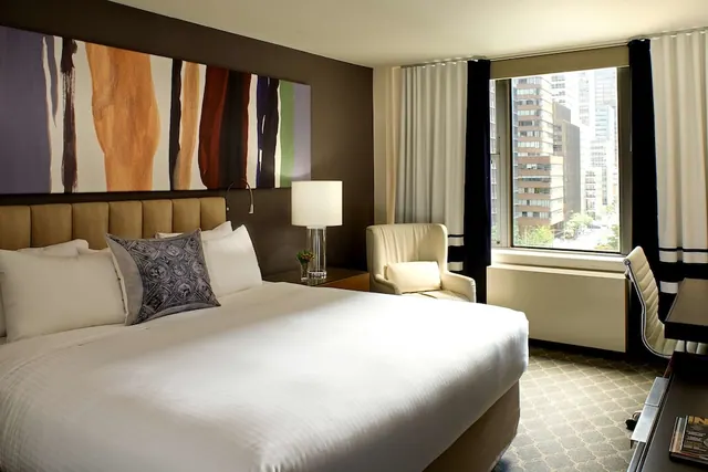 A hotel bedroom with a made king bed, an armchair by the window, and a desk on the right, with city buildings visible outside.