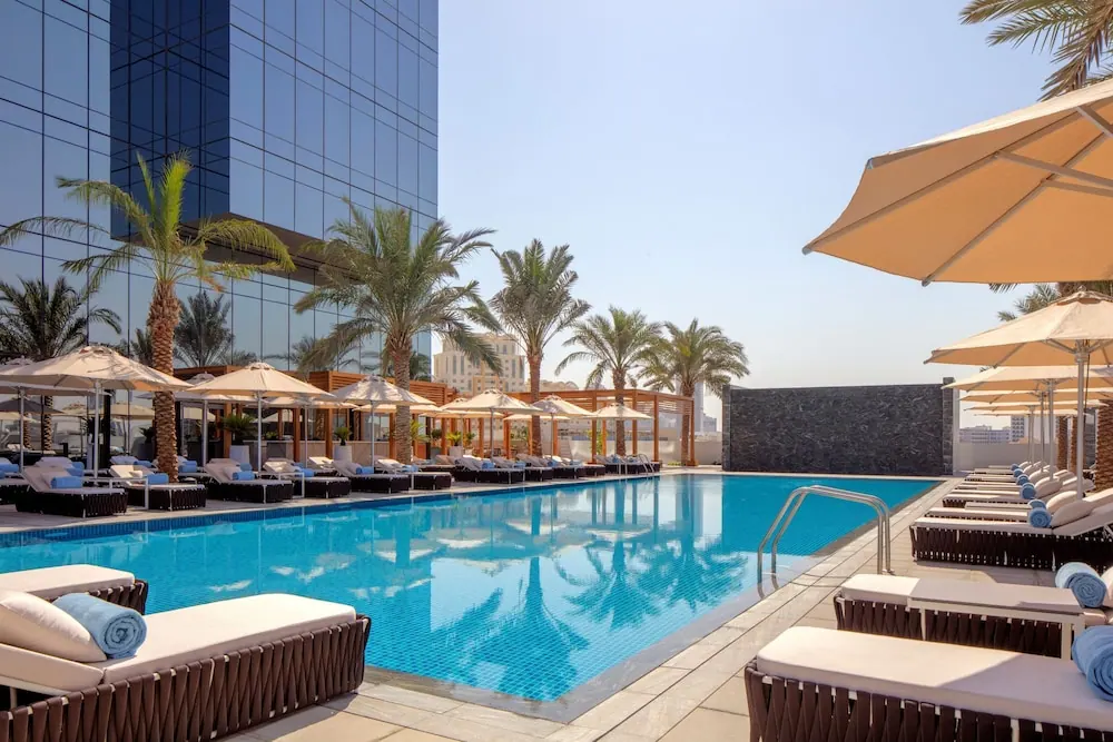 A rectangular outdoor swimming pool is surrounded by sun loungers, shade umbrellas and palm trees beside a glass-fronted building.