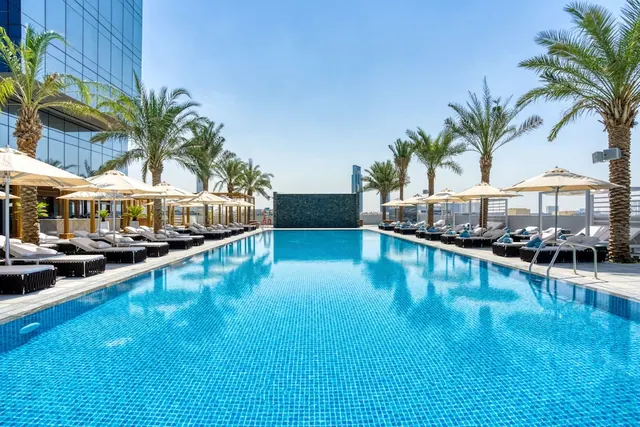 A rooftop swimming pool with blue mosaic tiles is lined with sun loungers and shade umbrellas, with palm trees and tall buildings beyond.