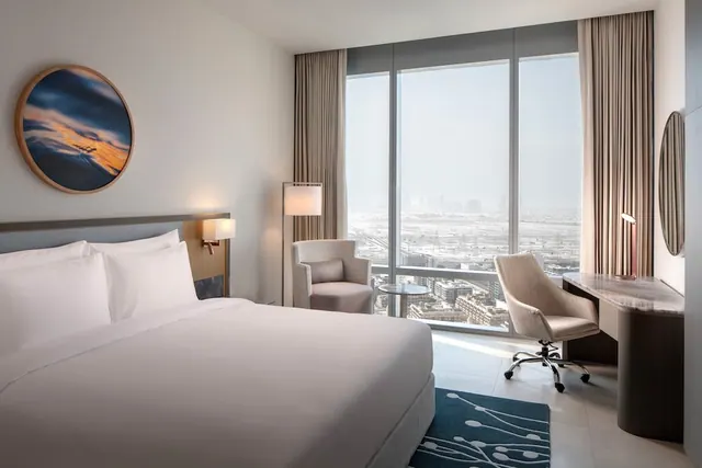 A bedroom with a king bed, an armchair, a desk and chair, and floor-to-ceiling windows looking out over a hazy city view.