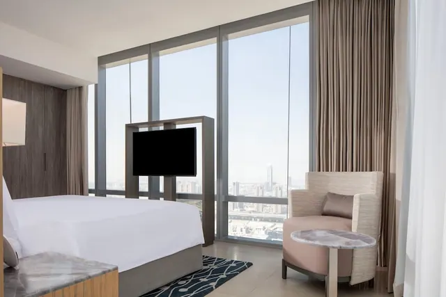 A bedroom with a made king bed, a television on a swivel stand, an armchair and a side table, with floor-to-ceiling windows overlooking a city skyline.