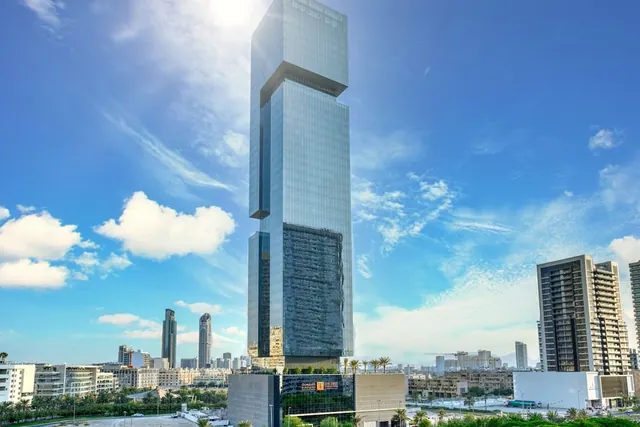 A glass-clad tower with an offset upper section rises above lower buildings under a blue sky, with other city buildings visible around it.