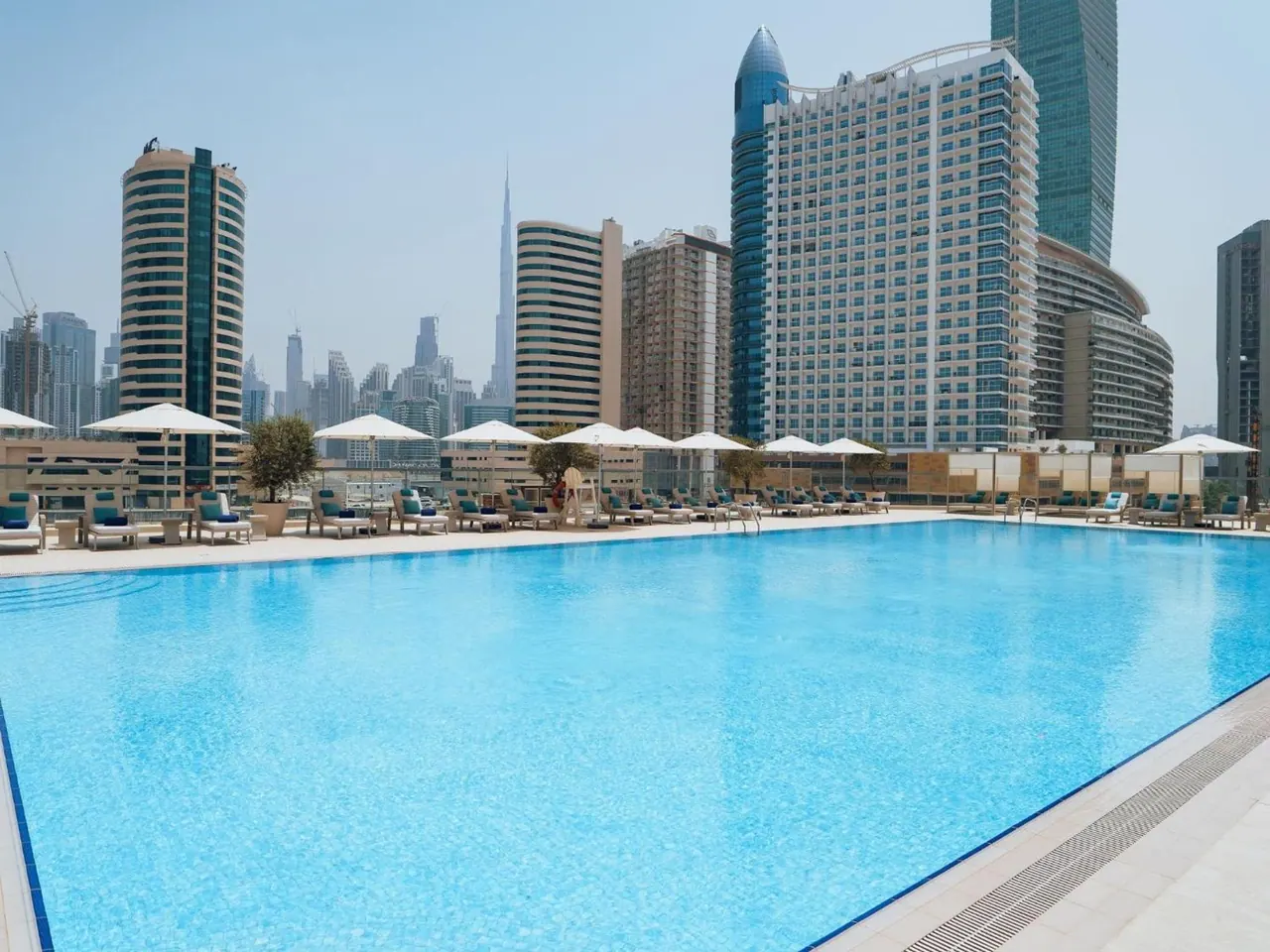 An outdoor swimming pool with white umbrellas and sun loungers in front of a city skyline with a tall spire tower in the background.