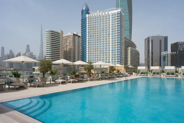 A large rooftop swimming pool with white parasol umbrellas and sun loungers, with high-rise towers and the Burj Khalifa in the background.