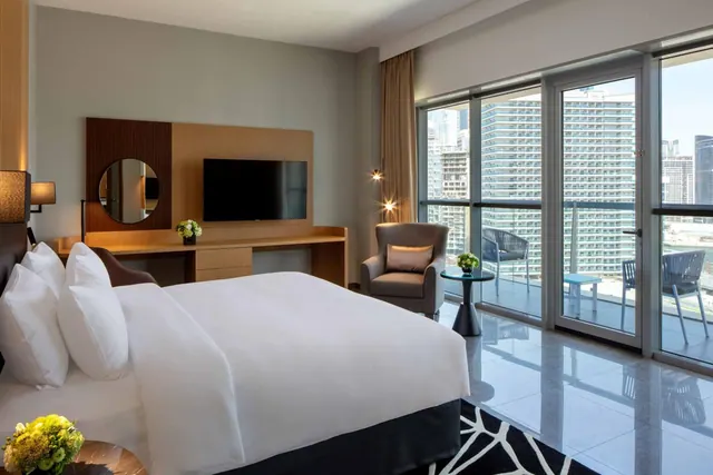 A bedroom with a king bed, a wall-mounted television, an armchair and a balcony with floor-to-ceiling glass doors overlooking high-rise towers.