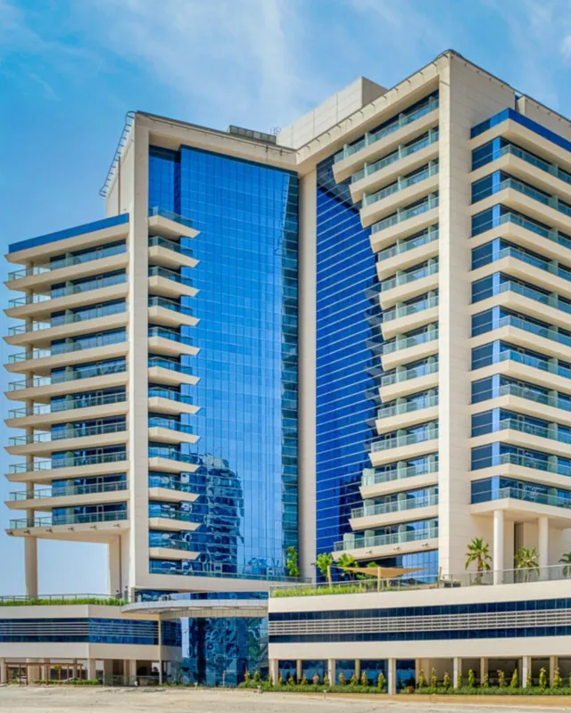Exterior view of a tall cream and blue glass tower with stacked balconies, palm trees and a low podium below under a clear blue sky.