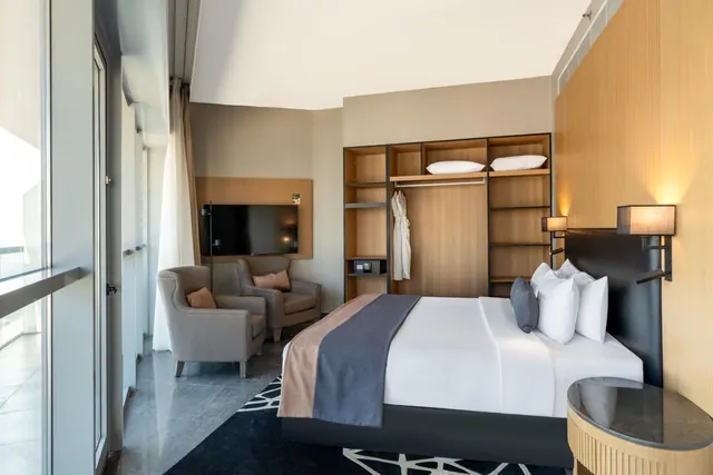 A bedroom with a king bed, two armchairs, a wall-mounted television, open wardrobe shelving and a floor-to-ceiling window on the left.