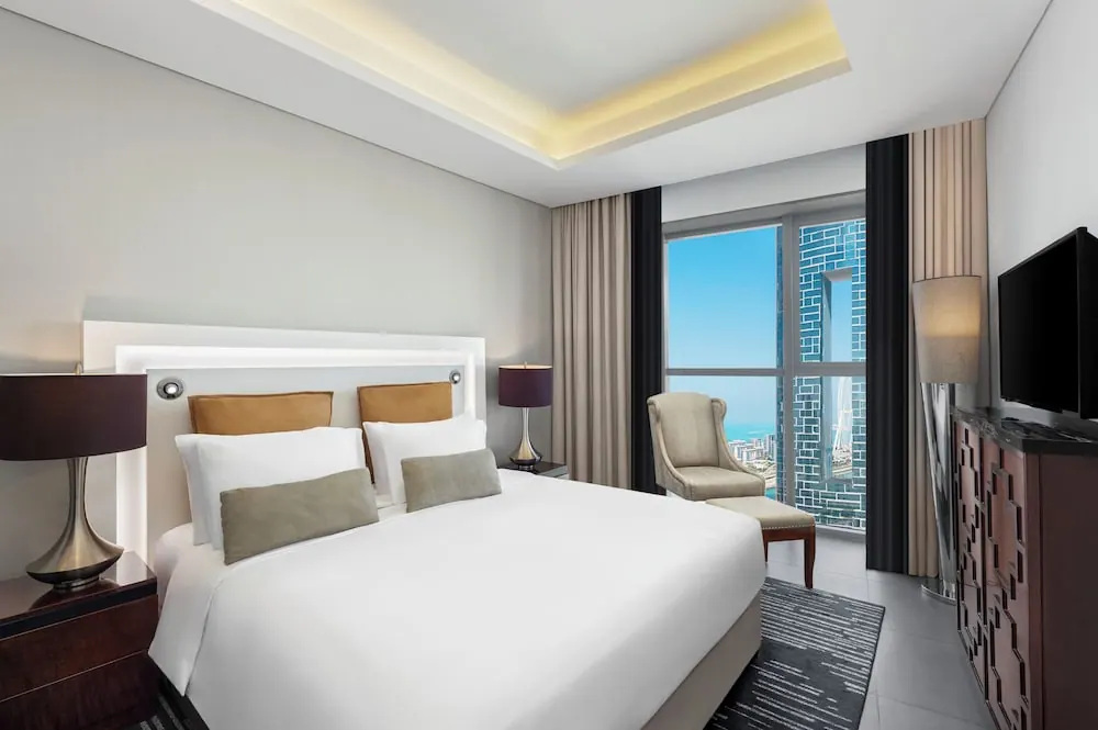 A bedroom with a made king bed, an armchair and ottoman beside a floor-to-ceiling window showing a strip of sea and buildings, and a wall-mounted television.