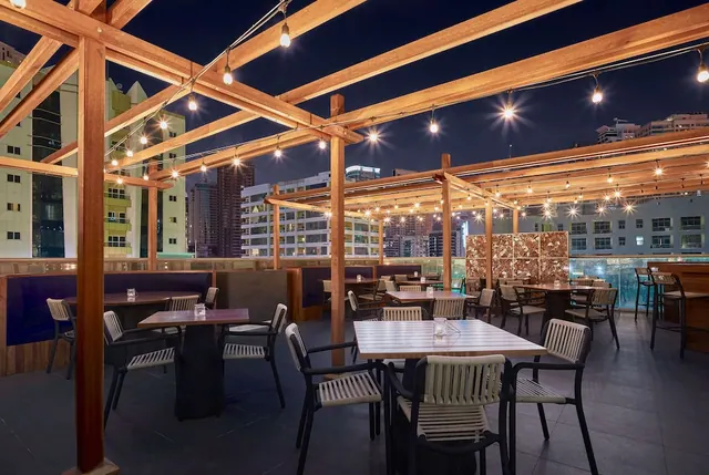 An outdoor rooftop dining area at night with a wooden pergola, string lights, tables and chairs, and city buildings beyond.