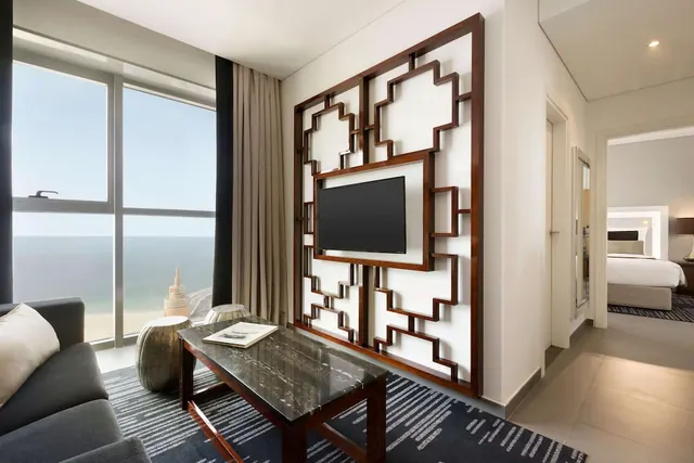 A living area with a grey sofa, a dark marble coffee table, a wall-mounted television in a carved wooden frame, and floor-to-ceiling windows looking out over the sea and a sandy beach.