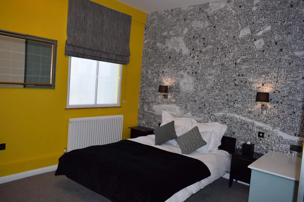A bedroom with a yellow wall, a grey patterned wallpaper feature wall, a double bed with a black throw, and wall lights on either side of the bed.