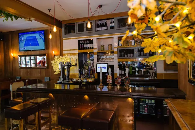 A pub-style bar with wood-panelled walls, beer taps, bar stools, a wall-mounted television showing tennis, and autumn leaves with fairy lights in the foreground.